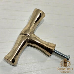 May include: A brass-colored cabinet pull handle in a T-shape, with a textured, organic design. The handle has a threaded bolt for attachment. The logo "QUENACRAFT ANTIQUE BRASS HARDWARE" is visible.