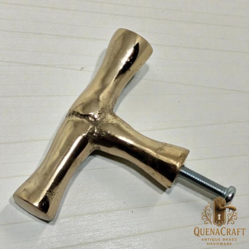 May include: A brass-colored cabinet pull handle in a T-shape, with a textured, organic design. The handle has a threaded bolt for attachment. The logo "QUENACRAFT ANTIQUE BRASS HARDWARE" is visible.