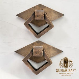 Może przedstawiać: Dwa antyczne mosiężne uchwyty do szuflad. Każdy uchwyt ma uchwyt w kształcie rombu w ramie w kształcie rombu, osadzony na tylnej płycie w kształcie rombu. Okucia mają rustykalny, postarzany wygląd.