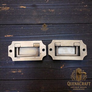 May include: Two antique brass drawer pulls with rectangular recessed handles. Each pull has a decorative, scalloped edge and two screw holes. The pulls are set against a dark wood background. The text "QUENACRAFT ANTIQUE BRASS HARDWARE" is visible.