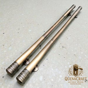 May include: Two antique brass door pulls with decorative end caps. The pulls are long and cylindrical, with a warm, golden-brown finish. The end caps feature intricate etched patterns. The hardware is designed for doors and cabinets.