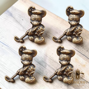 May include: Four brass Mickey Mouse cabinet knobs. Each knob depicts a seated figure with a hat, holding a golf club. The knobs are a warm, antique gold colour and are arranged on a light wood surface.