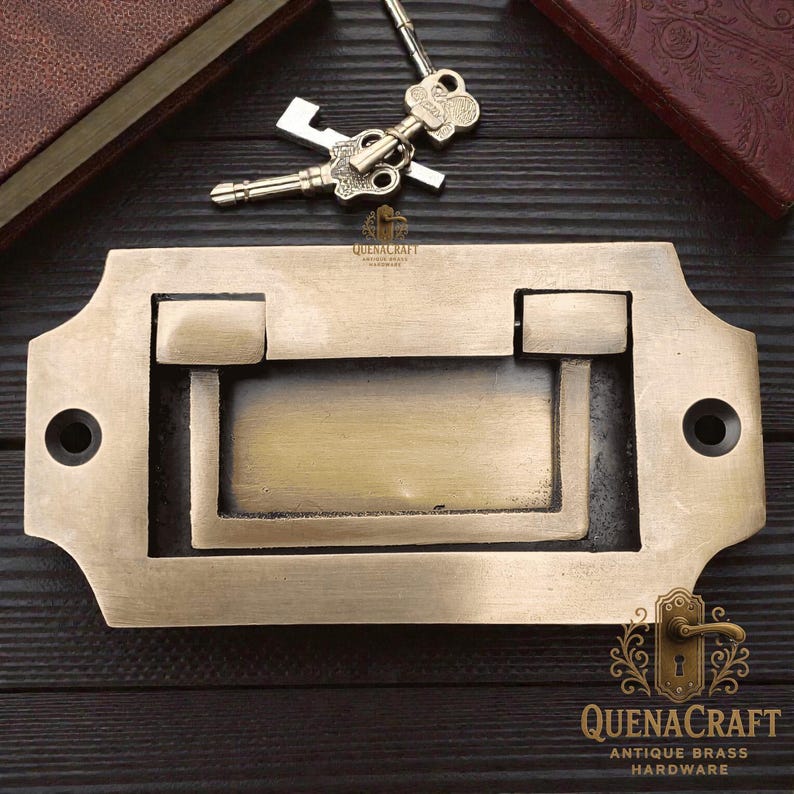 May include: Antique brass drawer pull with a rectangular design and a drop handle. The hardware features a vintage aesthetic, with two keys resting above. The product is from QuenaCraft Antique Brass Hardware.