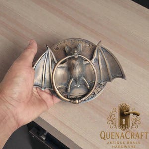 May include: Antique brass bat-shaped door knocker. The knocker features a detailed bat with outstretched wings and a circular handle. The design includes decorative elements and the text "QUENACRAFT ANTIQUE BRASS HARDWARE".