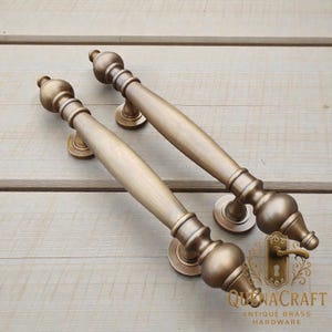May include: Two antique brass door handles with a classic design. Each handle features a long, cylindrical grip with decorative, rounded ends and circular mounting plates. The handles are displayed on a light wood surface.