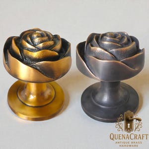 Op de afbeelding: Twee antieke messing kastknoppen in rozenvorm. De ene knop heeft een gouden afwerking, de andere een donker bronzen afwerking. Elke knop heeft een gedetailleerd rozenontwerp bovenop een ronde basis. De knoppen zijn van QuenaCraft Antique Brass Hardware.