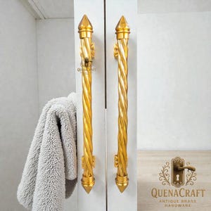 Puede incluir: Dos tiradores de armario dorados, de diseño retorcido, con extremos puntiagudos. Los tiradores están montados en una puerta de armario blanca. El texto "QUENACRAFT ANTIQUE BRASS HARDWARE" es visible.