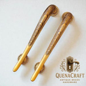 Puede incluir: Dos manijas de puerta de latón antiguo con un patrón detallado de vetas de madera. Las manijas son doradas con placas traseras circulares decorativas. La imagen incluye el logotipo de QuenaCraft Antique Brass Hardware.