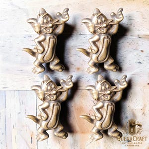 May include: Four antique brass cat-shaped cabinet pulls. Each pull features a smiling cat with arms raised. The pulls are a warm gold colour and are mounted on a light wood surface. The pulls are from QuenaCraft Antique Brass Hardware.