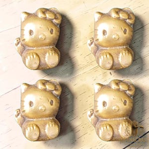 May include: Four gold-colored, cat-shaped figurines. Each figurine is in a seated position with one arm raised. The figurines have a bow on their head and are detailed with facial features and paws. The figurines are on a light brown surface.