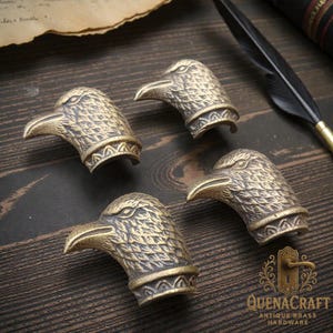 Pode incluir: Quatro puxadores de armário antigos em latão com cabeça de águia. Cada puxador apresenta detalhes de penas e uma faixa decorativa na base. Estão dispostos sobre uma superfície de madeira escura, com uma pena e um pergaminho ao fundo. O logótipo QuenaCraft é visível.