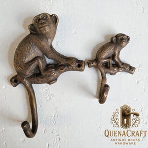 Puede incluir: Dos ganchos con forma de mono de color bronce. Los monos están posados en una rama, con el gancho que se extiende desde la cola del mono. Los ganchos son de latón y están diseñados para montaje en pared. El logotipo "QUENACRAFT ANTIQUE BRASS HARDWARE" está en la esquina inferior derecha.