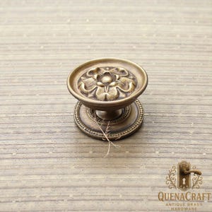 May include: Antique brass cabinet knob with a floral design. The knob features a round, raised face with a detailed flower pattern and a decorative base. The knob is made of brass and has a vintage aesthetic.