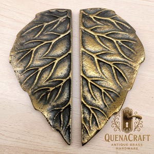 May include: Two antique brass leaf-shaped door handles. The handles have a detailed leaf vein design and a distressed finish. The handles are from QuenaCraft Antique Brass Hardware.