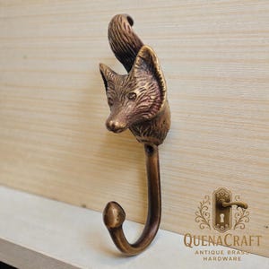 Puede incluir: Un gancho con forma de cabeza de zorro en color bronce, con un cuerpo curvado. La cabeza del zorro presenta detalles de pelaje y rasgos faciales. El gancho está montado sobre una superficie de madera clara. El logotipo "QUENACRAFT ANTIQUE BRASS HARDWARE" es visible.