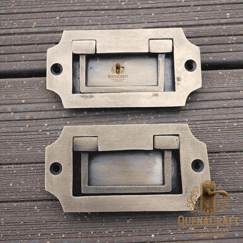 May include: Two antique-style drawer pulls in a muted silver-toned metal. Each pull has a rectangular recessed handle and decorative edges. The text "QUENACRAFT" is visible. Each pull has two screw holes for mounting.