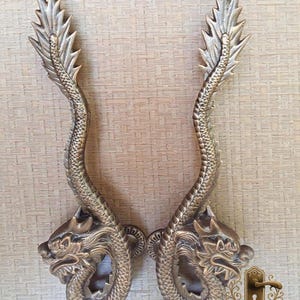 May include: Pair of antique brass dragon-shaped door handles. Each handle features a detailed dragon head and body, with a curved tail and ornate detailing. The handles are a warm, metallic color and are mounted on a light-colored surface.