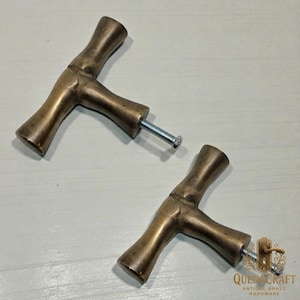 May include: Two antique brass cabinet handles with a T-shaped design. The handles have a warm, golden-brown colour and a textured surface. Each handle is attached to a silver screw. The handles are from QuenaCraft Antique Brass Hardware.