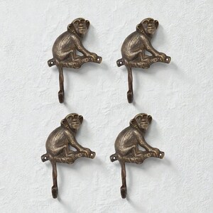 May include: Four bronze-effect monkey-shaped wall hooks. Each hook depicts a detailed monkey figure seated on a branch, with a curved hook below. The hooks are mounted on a textured, off-white surface.