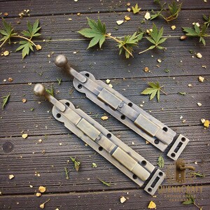 May include: Two antique brass sliding bolts with ball-shaped handles. The bolts are rectangular with screw holes and a sliding mechanism. The hardware is set on a dark wooden surface with scattered greenery.