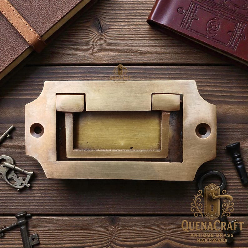 May include: A brass drawer pull with a rectangular design and a recessed handle. The hardware is surrounded by antique keys and leather-bound journals, suggesting a vintage aesthetic. The text "QUENACRAFT ANTIQUE BRASS HARDWARE" is visible.