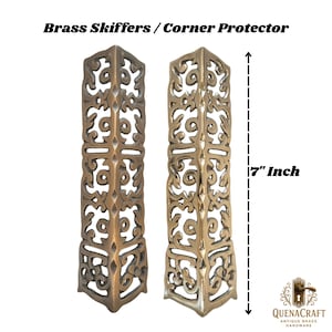 May include: Two brass corner protectors with an ornate, openwork design. Each piece is 7 inches tall. The text "Brass Skiffers / Corner Protector" is at the top. The QuenaCraft Antique Brass Hardware logo is at the bottom.