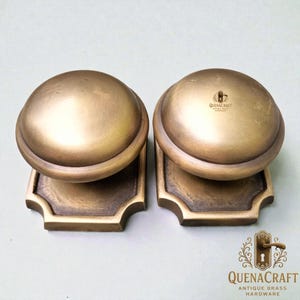 Könnte beinhalten: Zwei antike Messing-Türknöpfe mit rundem Design. Jeder Knopf sitzt auf einer quadratischen Basis mit dekorativen Kanten. Die Knöpfe haben einen warmen, goldenen Farbton. Der Text "QUENACRAFT ANTIQUE BRASS HARDWARE" ist enthalten.