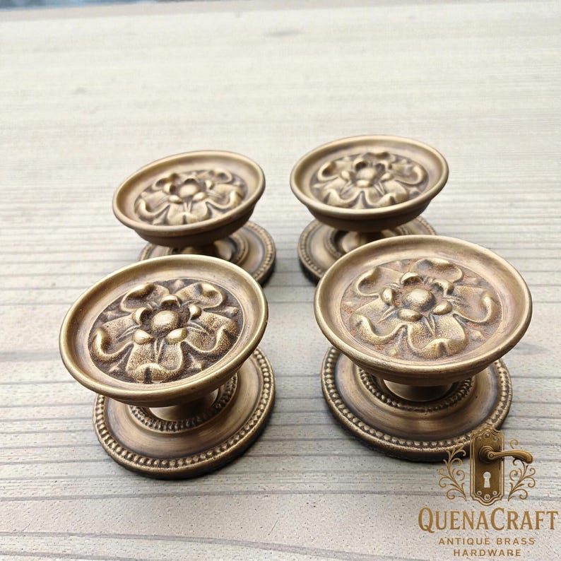 May include: Four antique brass cabinet knobs with a floral design. Each knob features a round, raised face with a detailed flower pattern and a beaded edge. The knobs are mounted on a circular base and are displayed on a light wood surface.