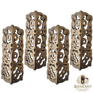 May include: Four antique brass corner brackets with intricate openwork designs. The brackets are a warm, aged bronze color, and the openwork creates a decorative pattern. The brackets are from QuenaCraft Antique Brass Hardware.