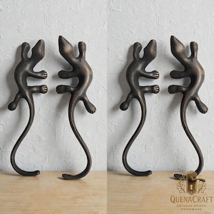 May include: Four bronze-colored lizard-shaped door handles. Each handle features a lizard design with a long, curved tail. The handles are displayed against a light-colored wall and a wooden surface. The text "QUENACRAFT ANTIQUE BRASS HARDWARE" is visible.