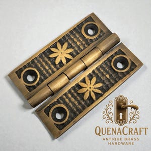 May include: Antique brass hinge featuring a floral pattern and grooved detailing. The hinge has two mounting holes on each side and a central pivot. The image also includes the text "QUENACRAFT ANTIQUE BRASS HARDWARE".