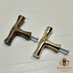 May include: Two brass T-shaped cabinet pulls with screws. One pull has a darker, antique finish, while the other is a bright gold color. The pulls are displayed on a light-colored surface.