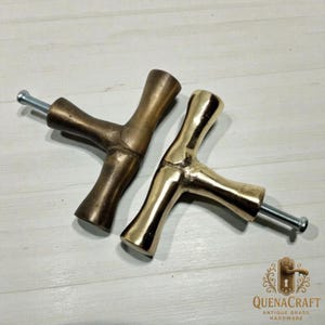 May include: Two brass cabinet pulls with a Y-shaped design. One pull has a darker, antique finish, while the other is polished gold. Each pull has a threaded screw for installation. The pulls are from QuenaCraft Antique Brass Hardware.