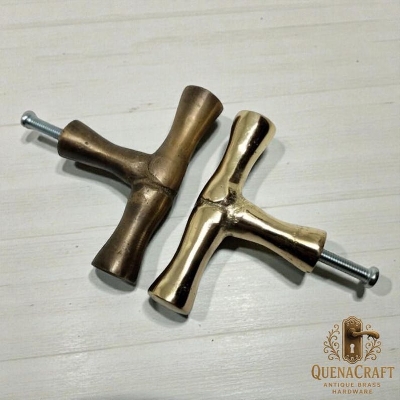 May include: Two brass cabinet pulls with a Y-shaped design. One pull has a darker, antique finish, while the other is polished gold. Each pull has a threaded screw for installation. The pulls are from QuenaCraft Antique Brass Hardware.