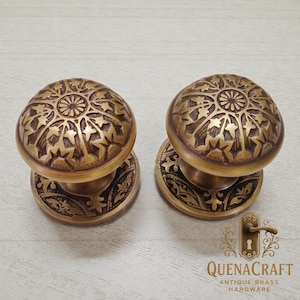 May include: Two antique brass door knobs with intricate floral and starburst patterns. The knobs are dome-shaped with matching circular backplates. The image also includes the text "QUENACRAFT ANTIQUE BRASS HARDWARE".