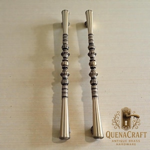May include: Two antique brass door handles with a decorative design. The handles have a long, slender shape with a series of decorative beads and rings. The handles are a warm, metallic color and are likely made of brass. The logo "QUENACRAFT ANTIQUE BRASS HARDWARE" is visible.