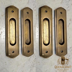 May include: Four antique brass recessed door pulls. Each pull has a rectangular shape with a rounded oval indentation in the centre. The pulls are a warm, aged brass colour, with a slight patina. The pulls are for antique brass hardware.
