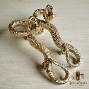 Może przedstawiać: Dwa mosiężne uchwyty do drzwi w kształcie węża. Uchwyty są w kolorze złotym ze szczegółowymi łuskami i otwartymi pyskami. Uchwyty są zamontowane na jasnej drewnianej powierzchni. Uchwyty pochodzą od QuenaCraft Antique Brass Hardware.