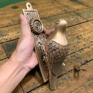 May include: A bronze-colored door handle with a bird design. The handle features a decorative backplate with a spiral design and a bird perched on the side. The handle is made of metal and has a vintage aesthetic.