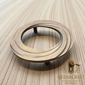 May include: Antique brass hardware ring with a spiral design. The circular ring features curved lines radiating from the centre. The hardware has three small legs and the text "QUENACRAFT ANTIQUE BRASS HARDWARE".