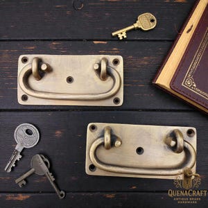 May include: Two antique brass drawer pulls with curved handles and rectangular backplates. Several vintage keys are scattered around the pulls, along with a leather-bound book. The hardware has a warm, aged finish, suggesting a vintage aesthetic.