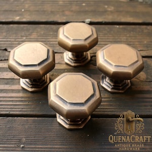 以下が含まれることがあります： 八角形のデザインの、アンティーク真鍮製のキャビネットノブ4つ。ノブは温かみのあるブロンズ色で、濃い木製の表面に置かれています。ノブはQuenaCraft Antique Brass Hardwareのものです。