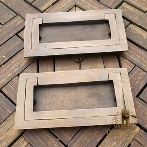 Puede incluir: Dos tiradores empotrados rectangulares de color bronce. Cada tirador tiene un marco rectangular con un centro empotrado. Los tiradores están diseñados para quedar al ras del frente del cajón. Los tiradores son de metal y tienen un aspecto vintage o antiguo.