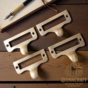 May include: Four antique brass label holders with a rectangular frame and a rounded handle. They are arranged on a wooden surface with a notebook and pen in the background. The logo "QUENACRAFT ANTIQUE BRASS HARDWARE" is visible.