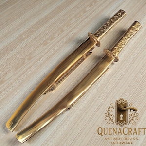 Puede incluir: Dos espadas decorativas doradas con empuñaduras y vainas ornamentadas. Se exhiben sobre una superficie de madera clara. La imagen incluye el texto "QUENACRAFT ANTIQUE BRASS HARDWARE".