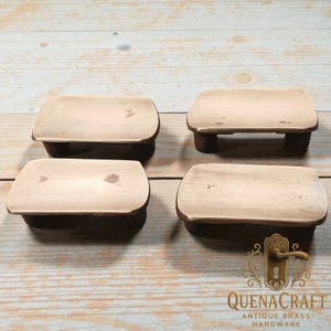 Könnte beinhalten: Vier rechteckige Möbelgriffe aus Holz im antiken Stil mit einem Used-Look. Die Griffe haben eine hellbeige Oberseite und dunkelbraune Seiten und Beine. Das Bild enthält das Logo von QuenaCraft Antique Brass Hardware.