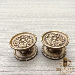 May include: Two antique brass hardware knobs with a floral design. The knobs have a round base with a beaded edge and a raised floral pattern on the top. The knobs are a warm, golden color.