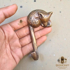 Könnte beinhalten: Ein messingfarbener, fuchsformiger Haken mit geschwungenem Design. Der Fuchs befindet sich in einer zusammengerollten Position, wobei sein Kopf auf seinen Pfoten ruht. Der Haken hat eine warme, antike Messingfarbe. Der Haken wird in einer Hand gehalten.