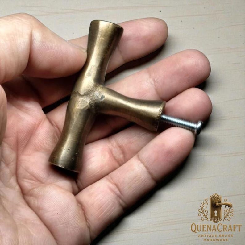 May include: A brass T-shaped cabinet pull with a screw attached. The hardware has a vintage aesthetic and a warm, golden hue. The product is from QuenaCraft Antique Brass Hardware.