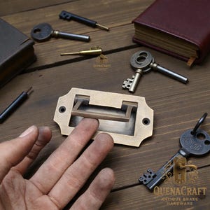 May include: A selection of antique brass hardware, including keys and a drawer pull. The drawer pull is rectangular with a recessed handle. Several keys of varying sizes and designs are scattered around the pull. The image is set on a wooden surface.